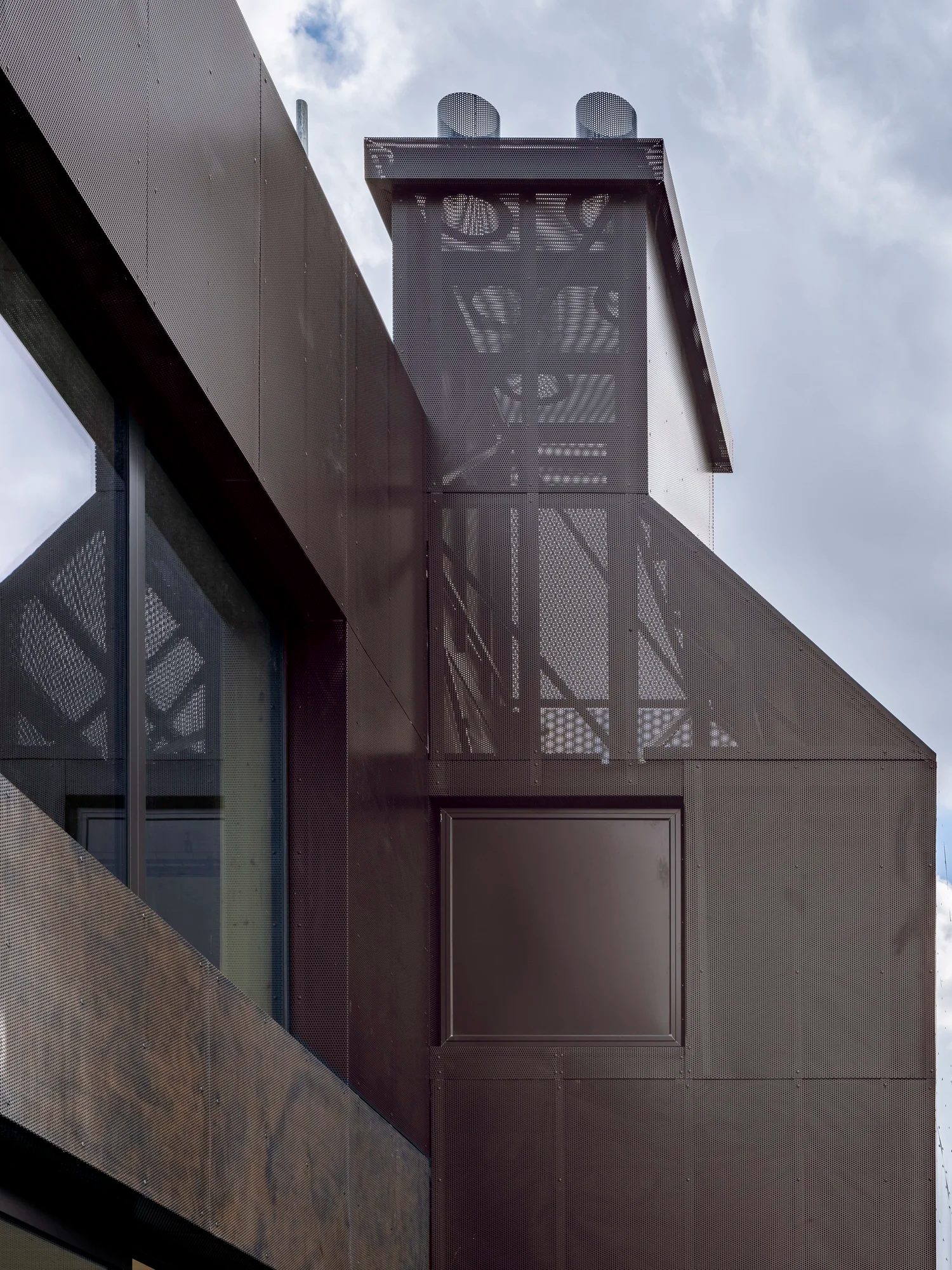Roof-level chimney form clad in folded perforated metal panels beside flat dark cladding and a glazed storey
