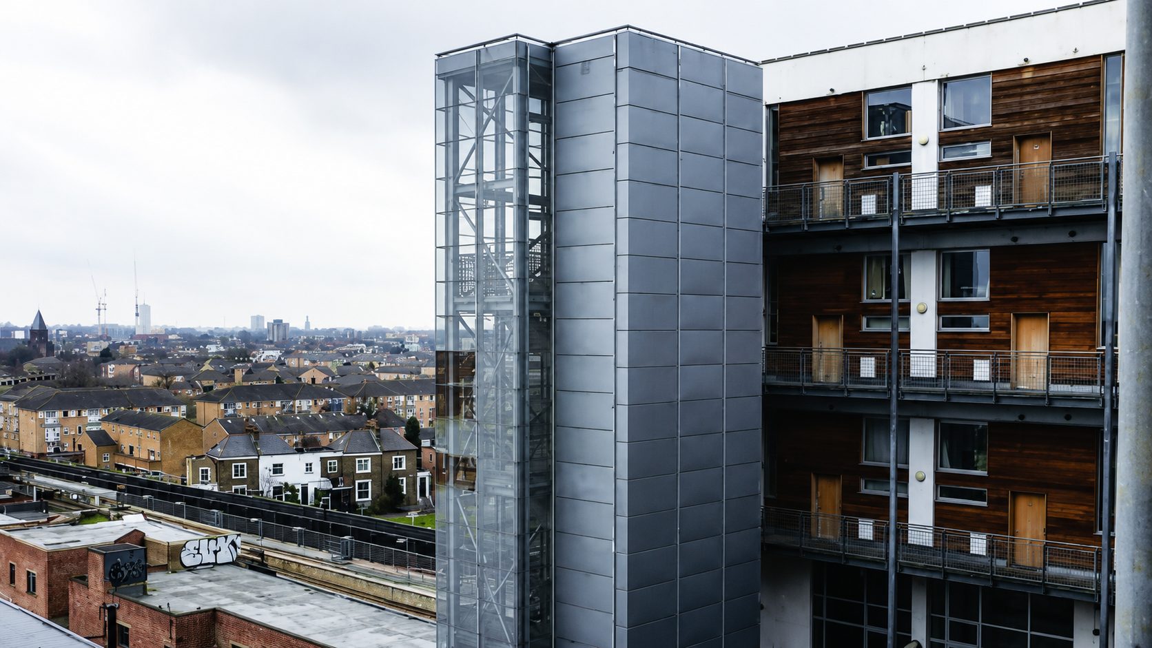 A glazed lift shaft and a metal-panel-clad tower at Ability Plaza, with the surrounding Haggerston rooftops beyond