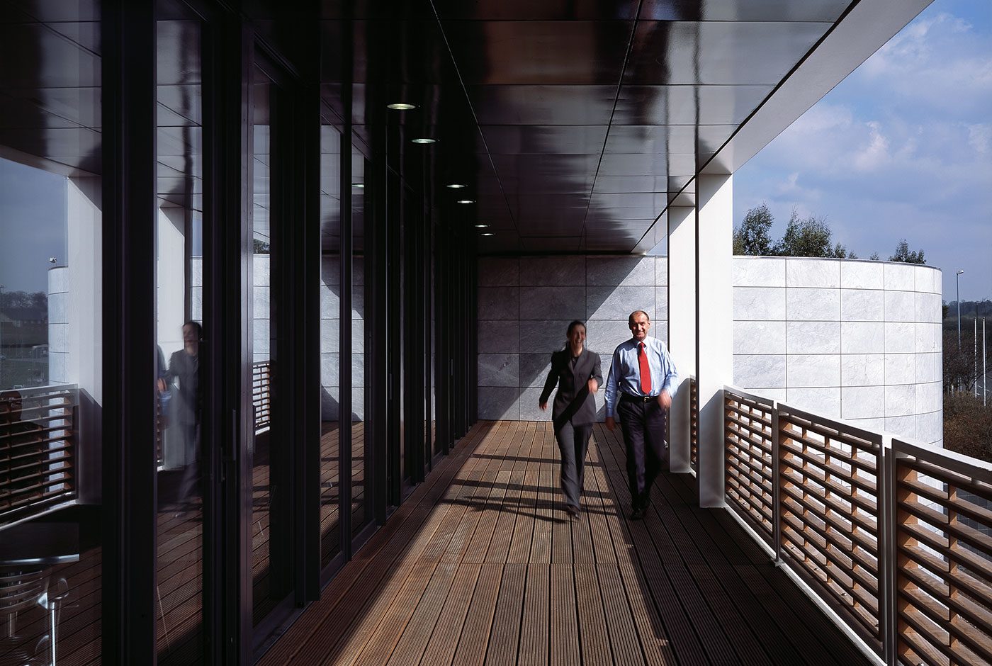 Two people walking along a timber-decked balcony beneath the cantilevered white soffit of Ashfield House's extension, a curved stone-clad drum to the right