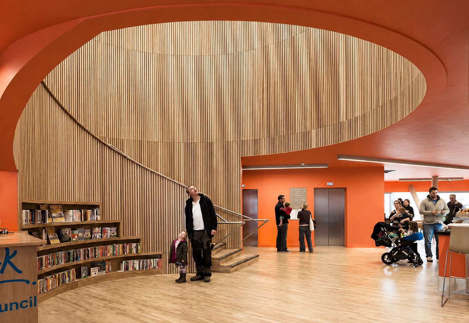 Canada Water Library's entrance hall, the timber-ribbed stair drum rising through an oval opening in the orange ceiling, visitors below