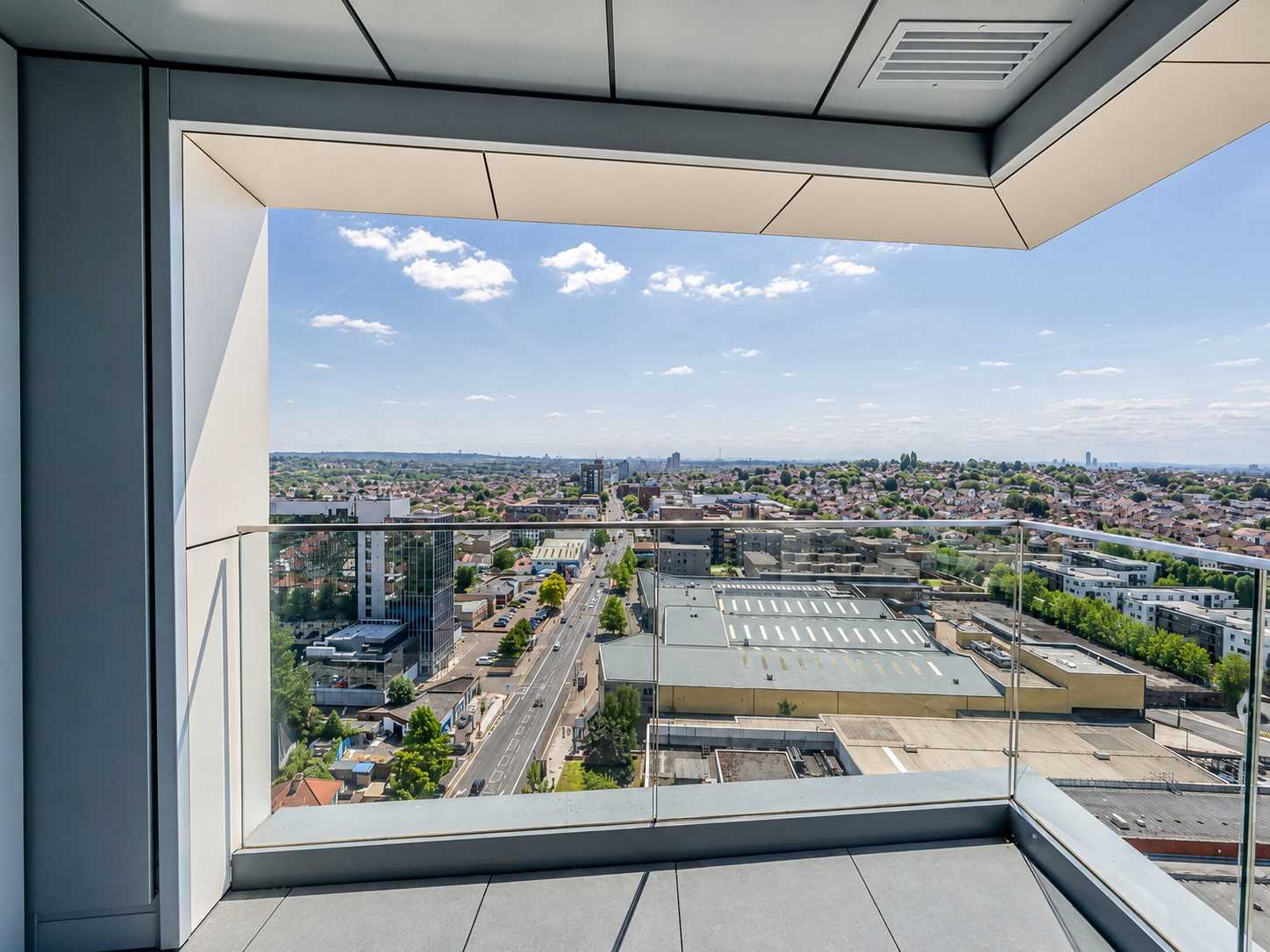 A corner balcony near the top of Everly House, its glazed balustrade and soffit framing a view north over Colindale and the London suburbs beyond