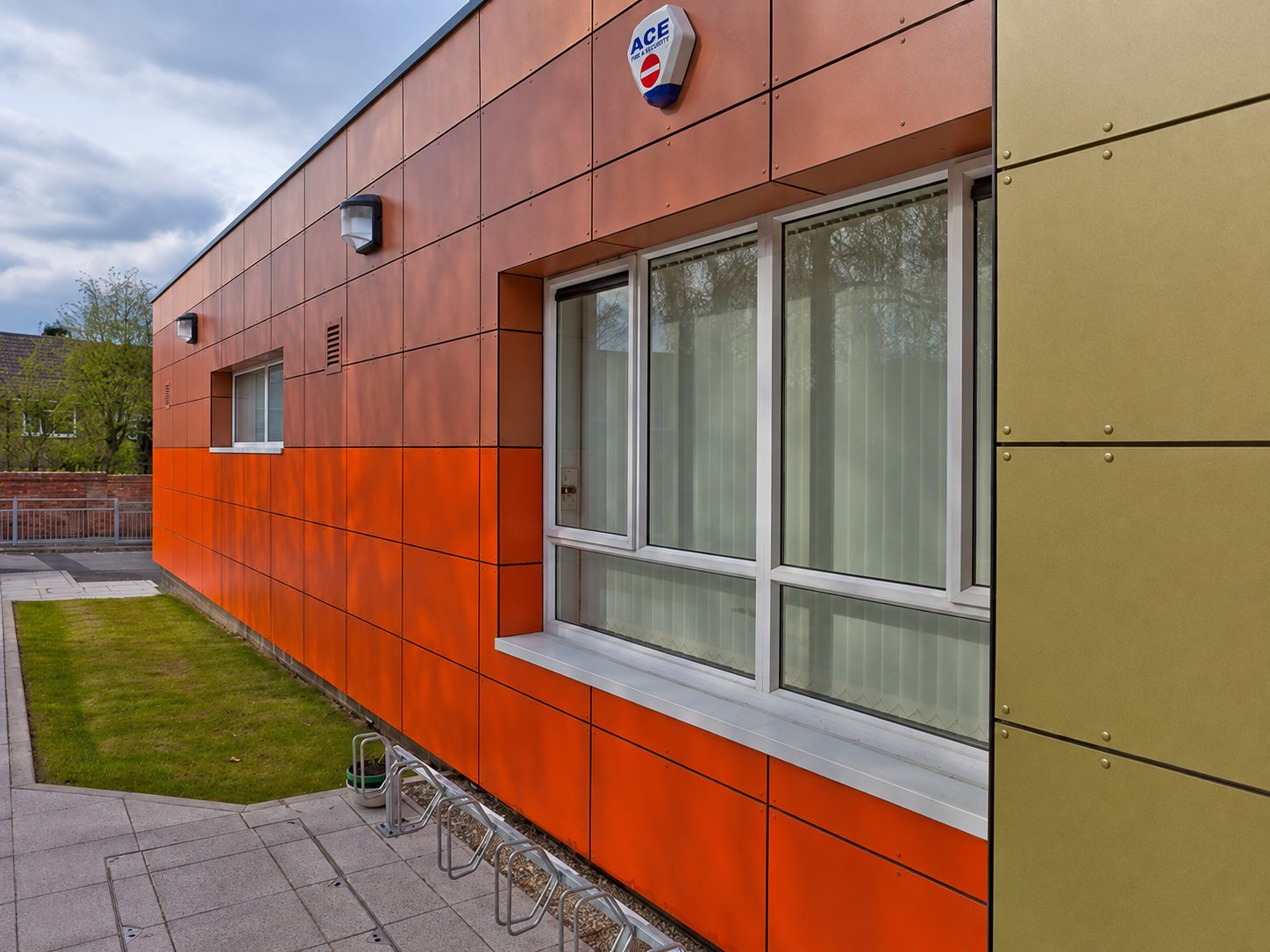 Orange cladding panels turning the corner of Hawes Down Centre past a window and a fire alarm bellbox, with grass and paving in the foreground