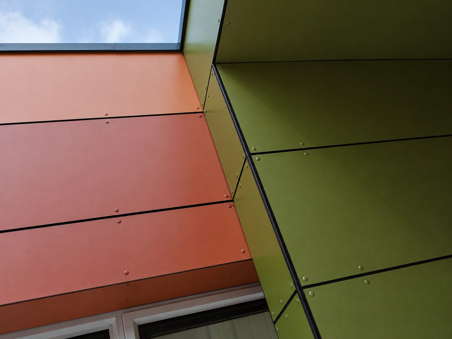 The corner where orange cladding panels meet the olive-green return at Hawes Down Centre, seen from below against the sky
