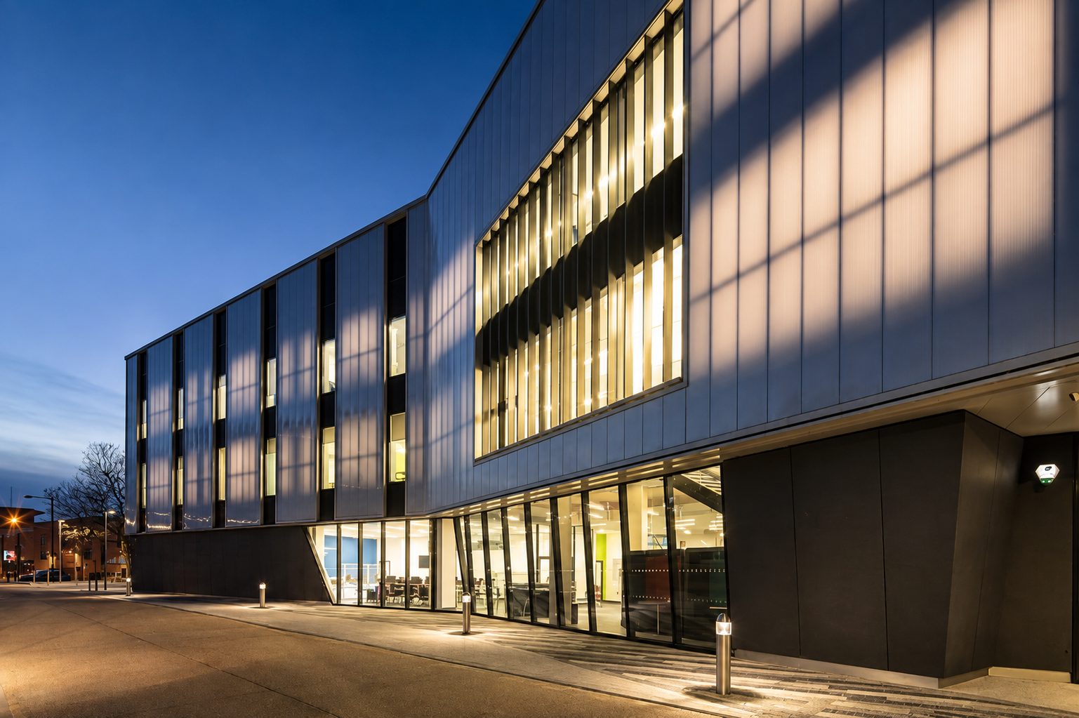 Close view of the Centre for Digital Technologies' illuminated vertical glazed bays at dusk, set within pale ribbed cladding panels