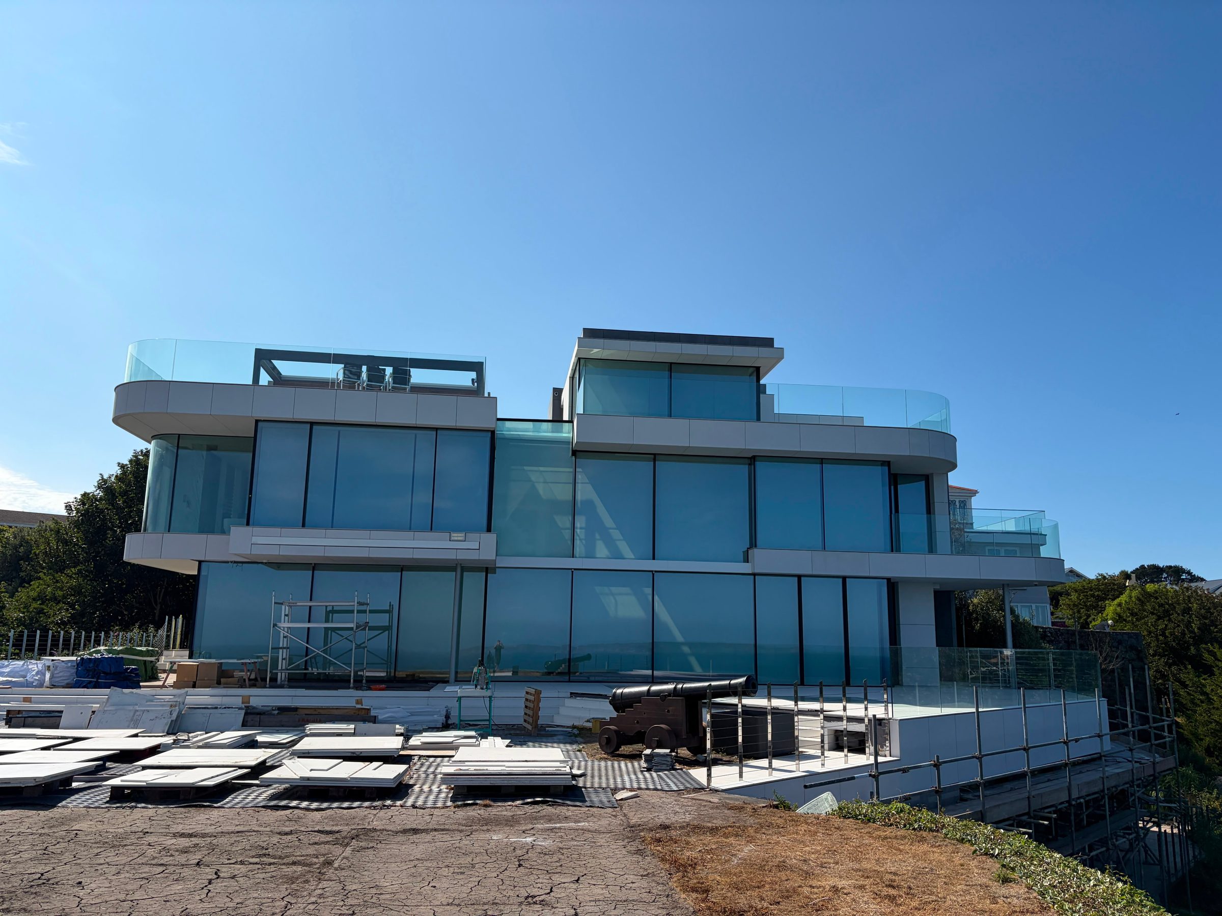 The Lighthouse's curved sea-facing elevation with glazed balconies and pale engineered stone panels, seen from the garden with construction materials still on site