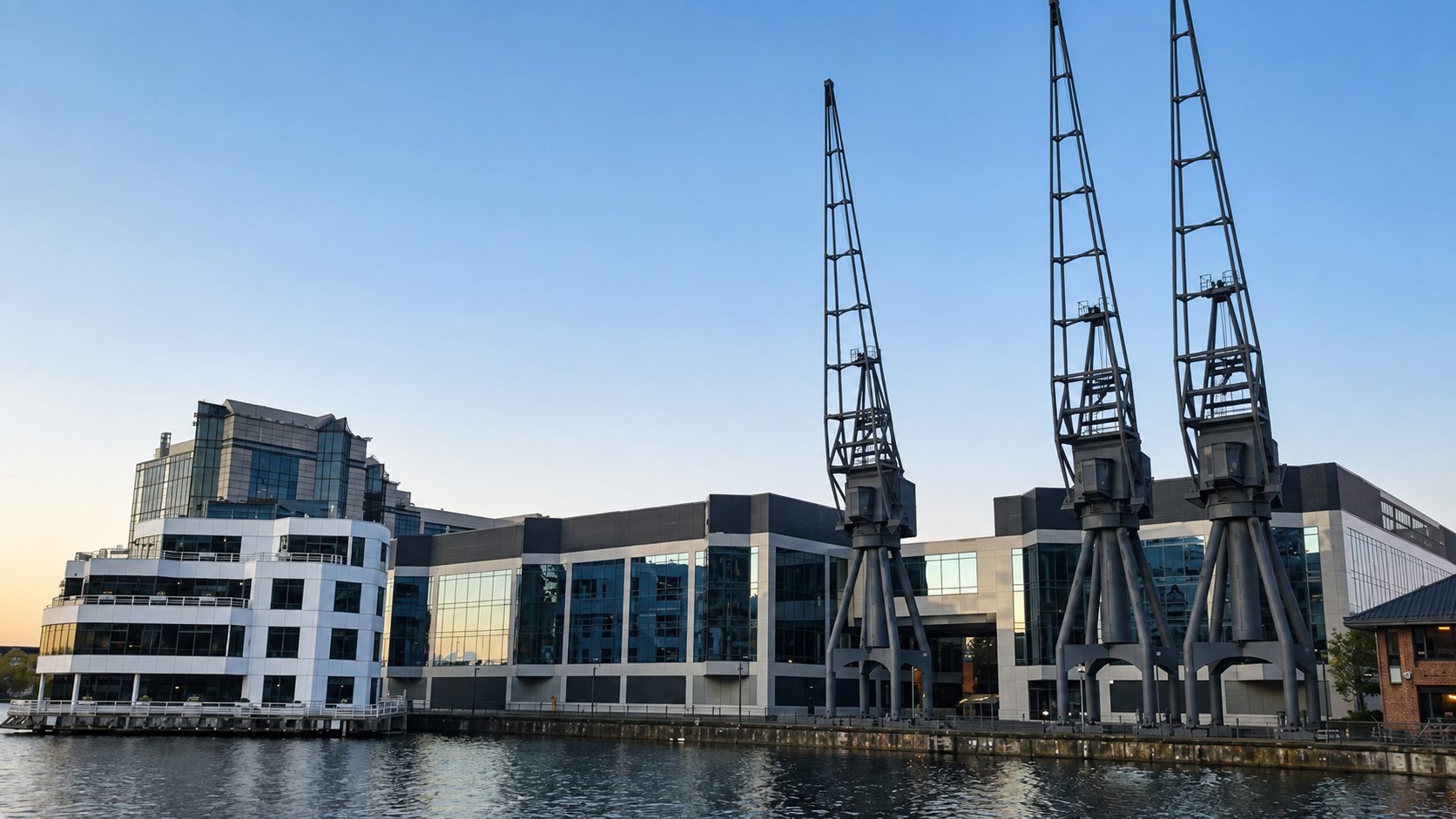 Glazed and pale-panelled dockside buildings behind three preserved cranes on the Isle of Dogs, the waterside setting of The Point