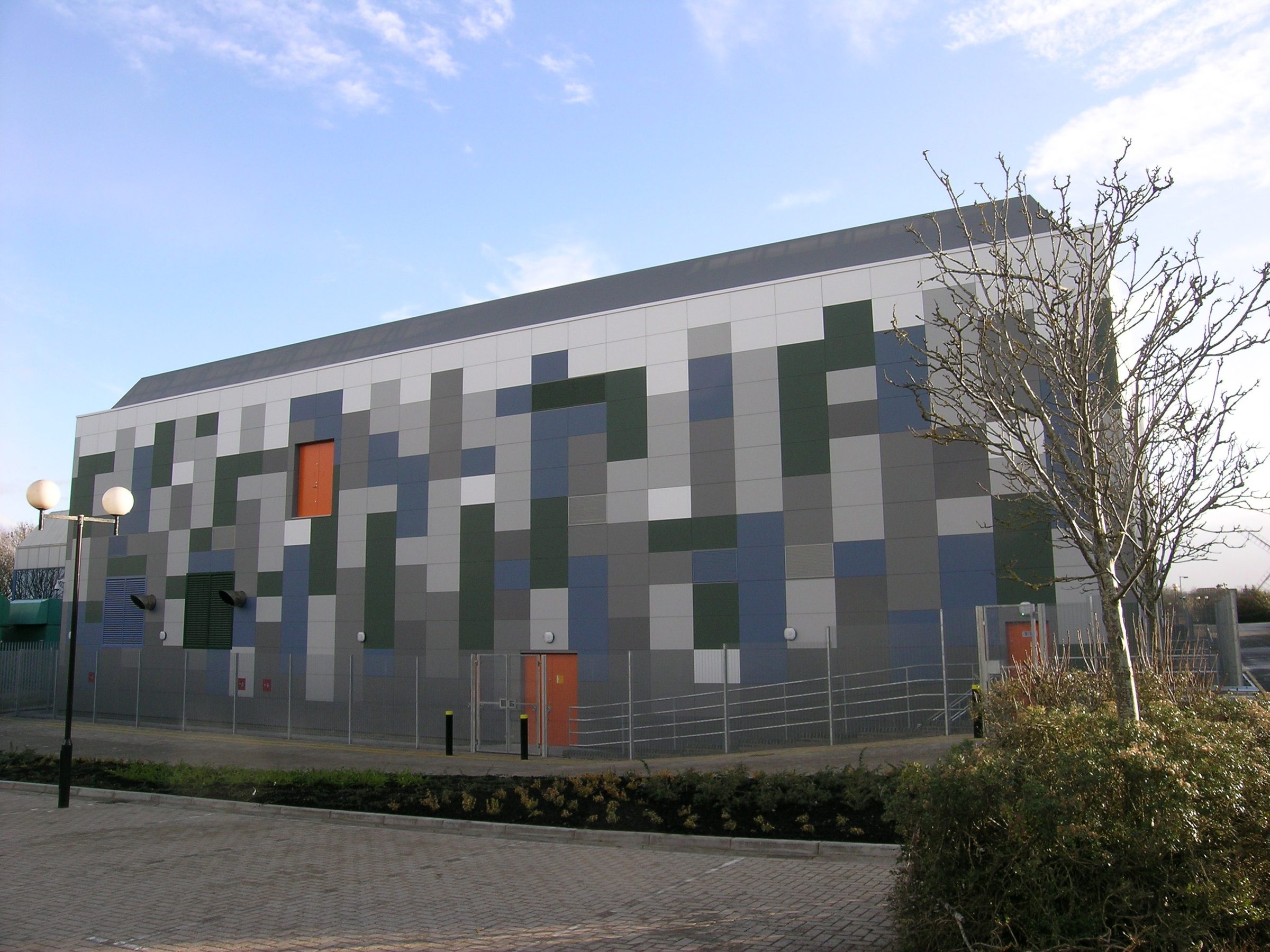 Building at Vodafone's Swindon data centre seen from its opposite corner, graduated blue-grey and green Eternit Pictura cladding panels around two orange doors, a bare tree in the foreground