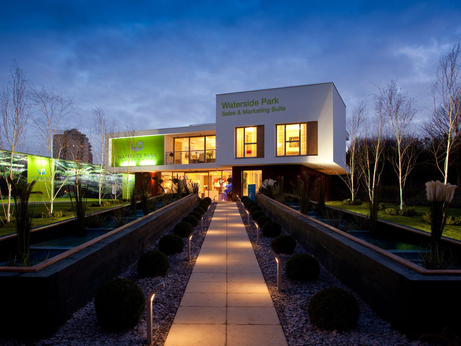 Waterside Park's Sales & Marketing Suite illuminated at dusk, its lit approach walkway flanked by reflecting pools