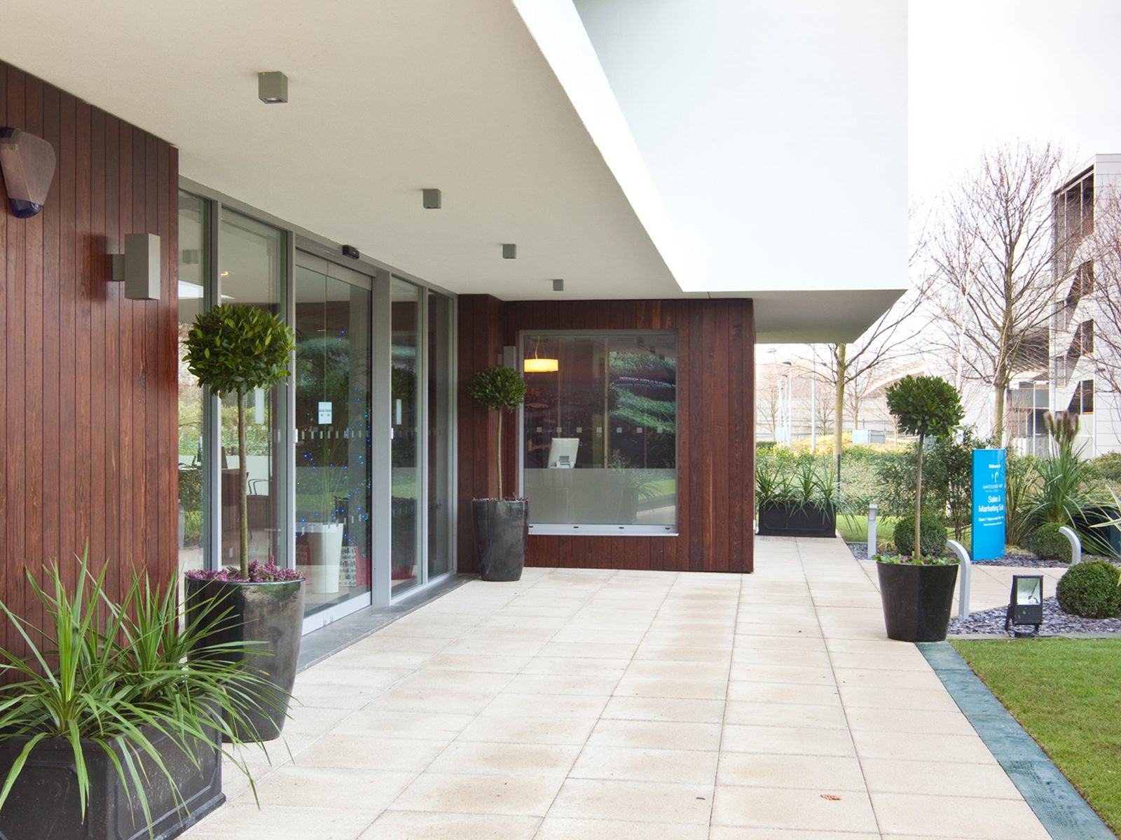 Dark stained vertical timber cladding under the entrance canopy at Waterside Park's Sales & Marketing Suite, potted bay trees either side of the glazed doors