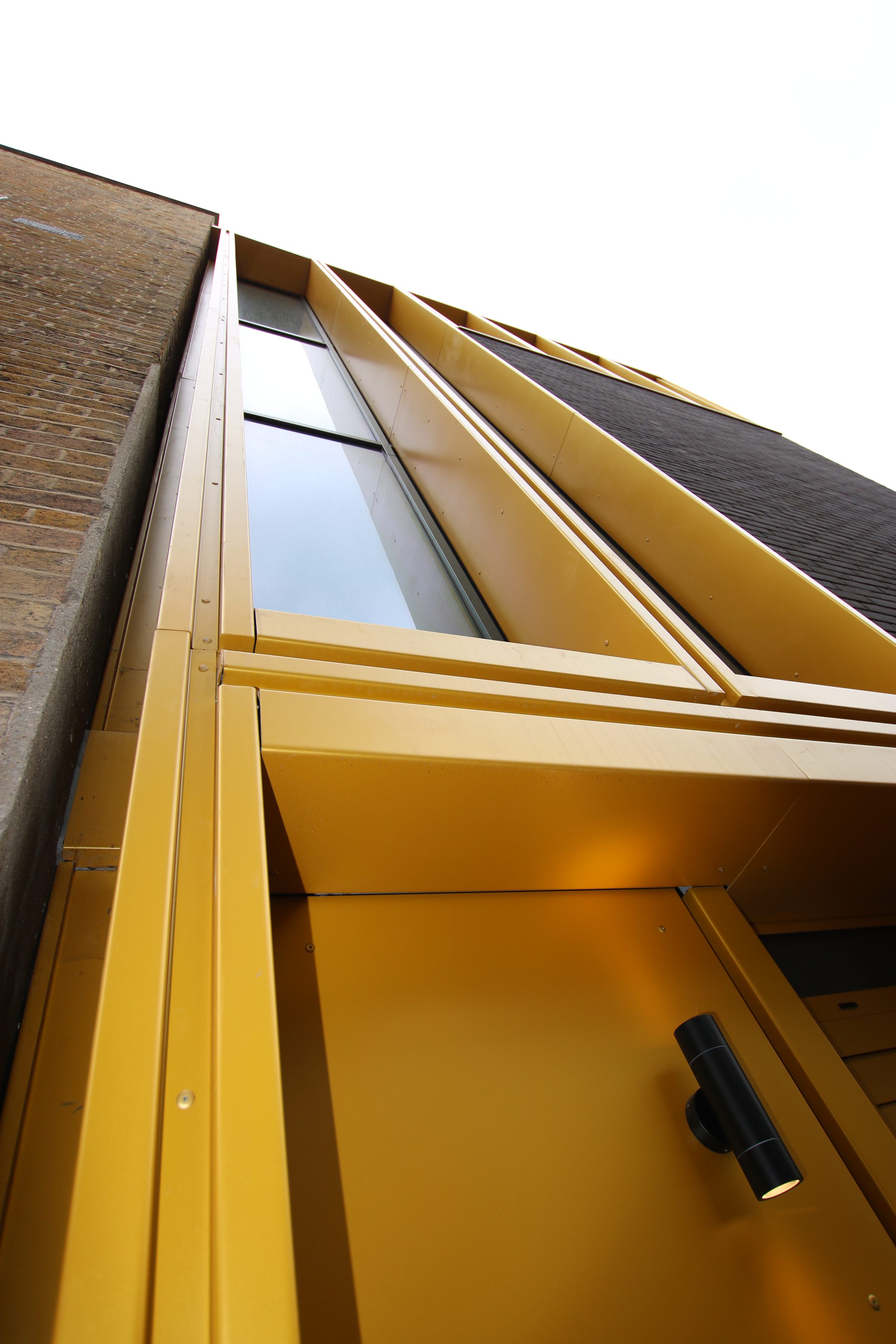 Looking up the gold aluminium rainscreen fins, reveals and soffit beside the old brick wall at William Gaitskell House, Rotherhithe